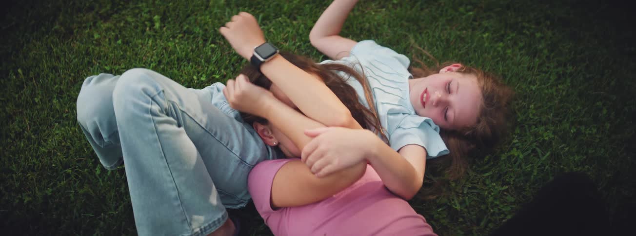 Two young sisters playfully wrestle and laugh while lying on the green grass in a park