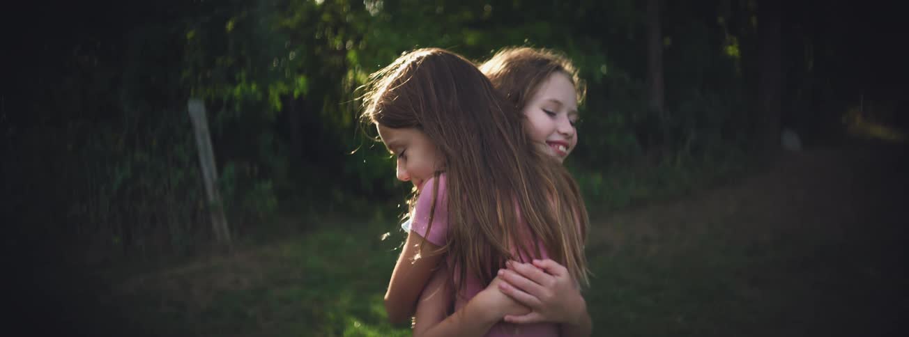 Two smiling girls embrace in a slow-motion, heartwarming hug outdoors during golden hour