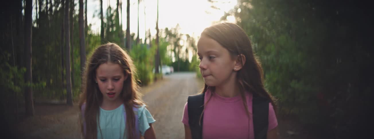 Close-up slow motion shot of two young girls with backpacks walking and talking on a wooded path at sunset