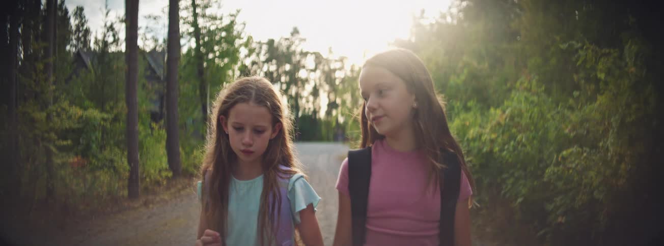 Two young sisters with backpacks walk home from school together, talking and smiling as they stroll down a sunlit, tree-lined road