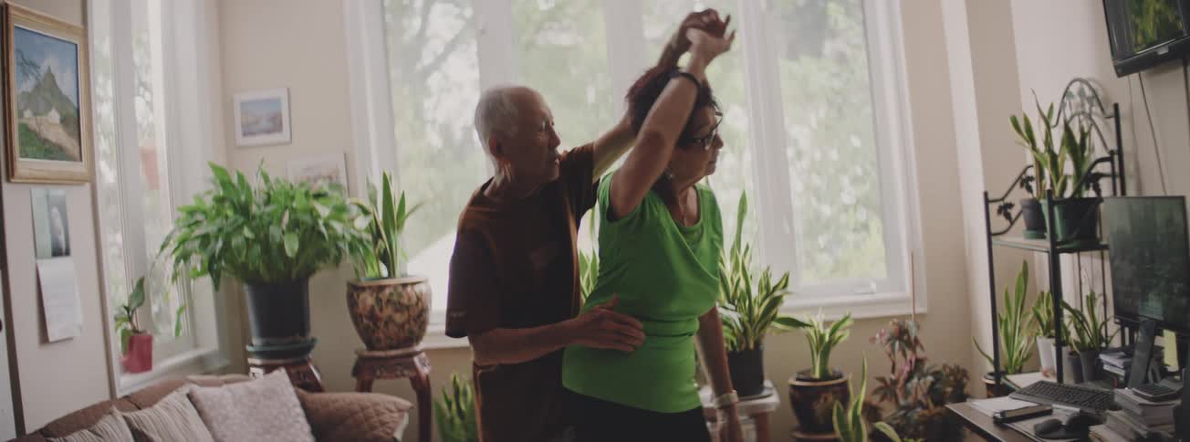 A loving senior couple shares a slow dance in their sun-drenched living room surrounded by houseplants