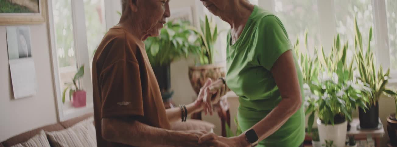 A senior Asian man and his wife share a tender slow dance in their plant-filled living room