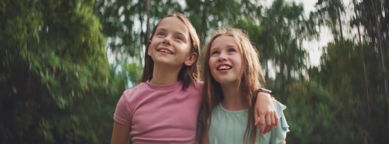 Two amazed young sisters look up at the sky with expressions of wonder and joy while standing together in a lush green park