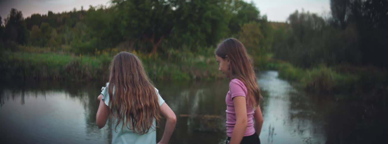 In slow motion, a young girl blows dandelion seeds over a tranquil river as her friend watches