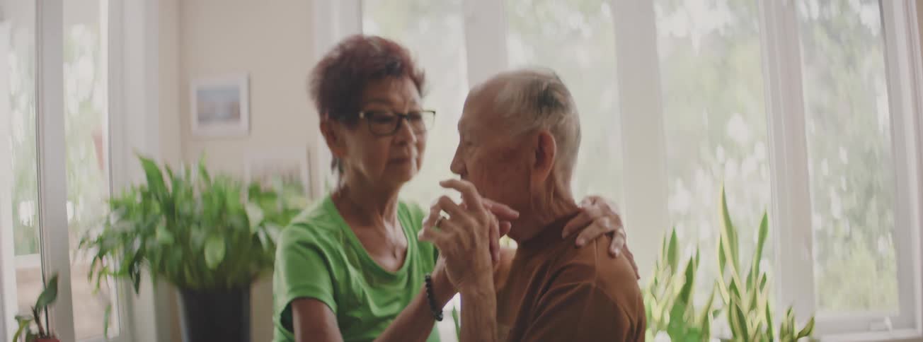 An elderly Asian couple shares a tender moment, slow dancing together in their sunlit home