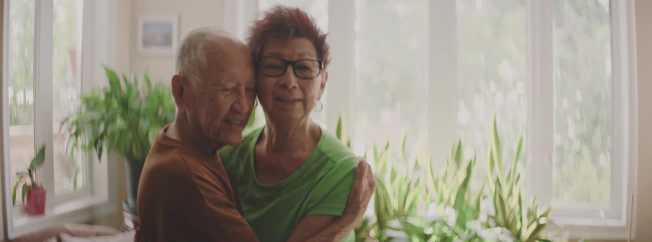 A joyful elderly Asian couple share a heartfelt laugh before embracing tenderly by a sunlit window in their home