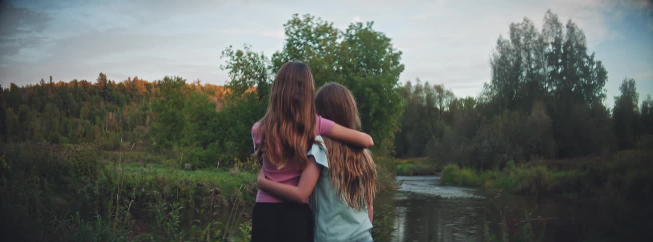 From behind, two young sisters or friends with long hair stand arm-in-arm, sharing a quiet moment by a tranquil river in nature