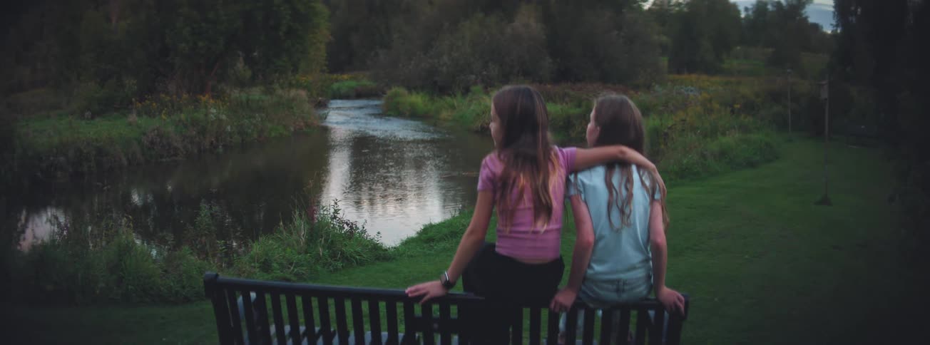 Two young sisters share a quiet moment with their arms around each other on a park bench overlooking a stream