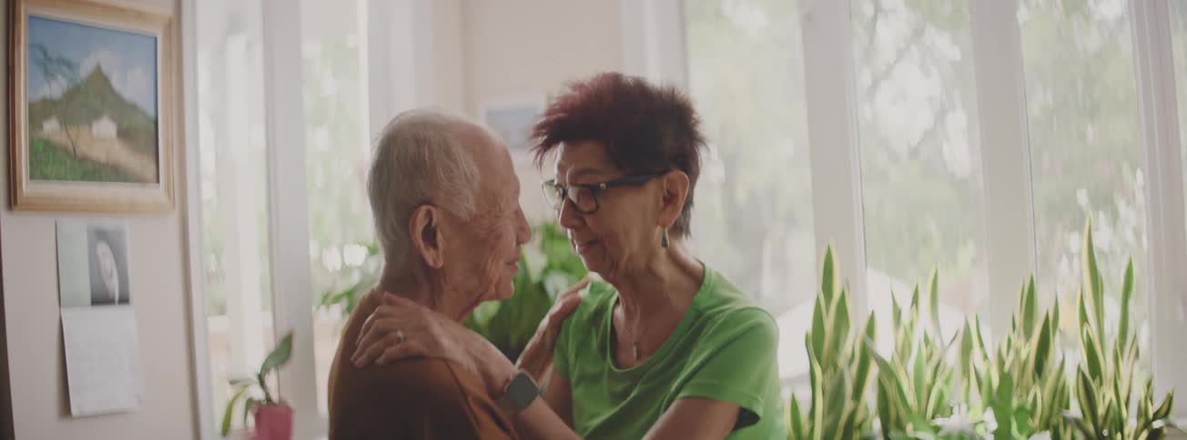 An elderly couple shares a loving and tender moment, slow dancing together in their sunlit, plant-filled living room
