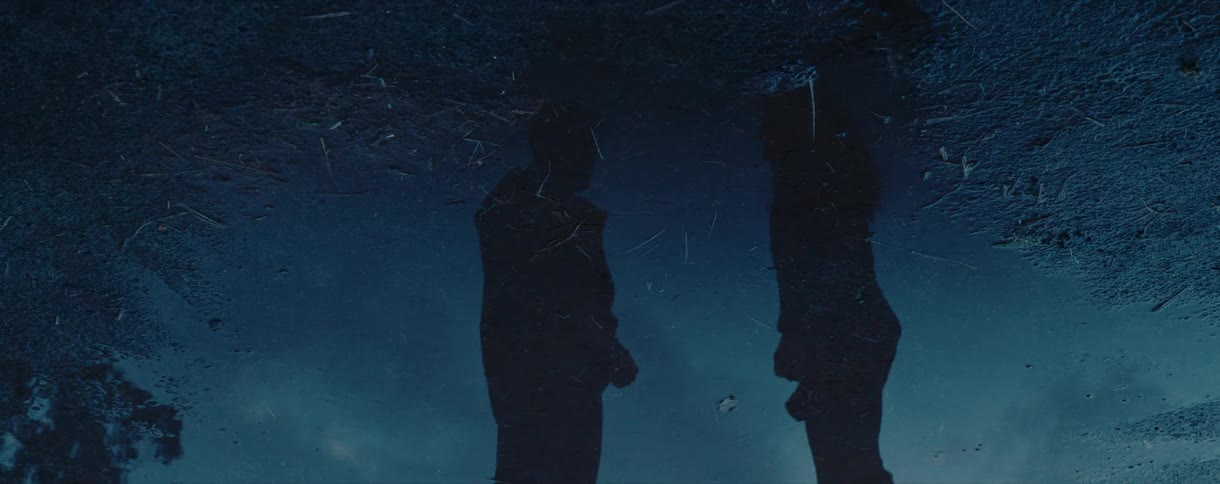 The mysterious silhouettes of two people are reflected in a dark puddle as gentle rain falls