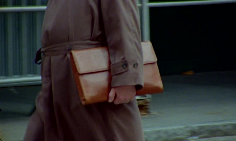 A man in a brown trench coat carrying a leather portfolio walks down a city street