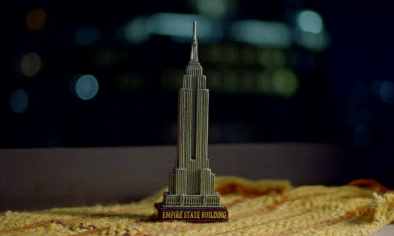 A miniature souvenir replica of the Empire State Building sits on a ledge with bokeh city lights in the background at night