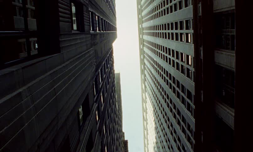 A low-angle worm's-eye view looking up between two imposing skyscrapers towards a bright sky