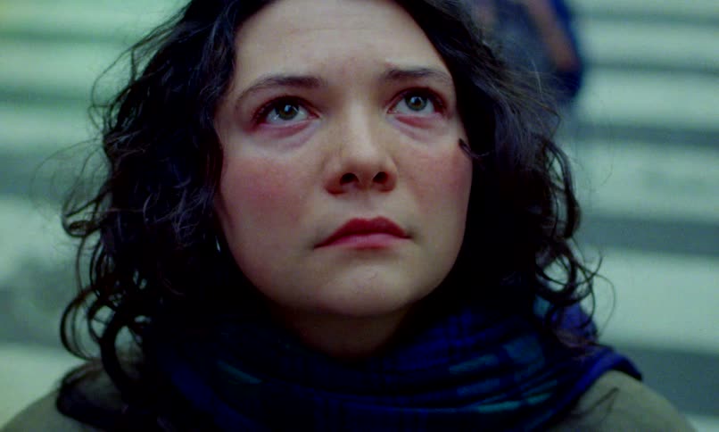 Close-up of a somber woman with dark curly hair and teary eyes looking upward in a busy public space