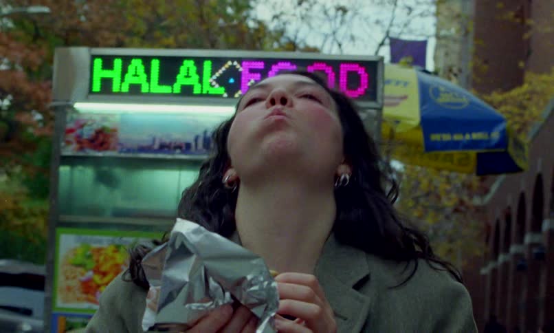 A woman with dark curly hair enthusiastically eats street food from a Halal food cart on a city sidewalk