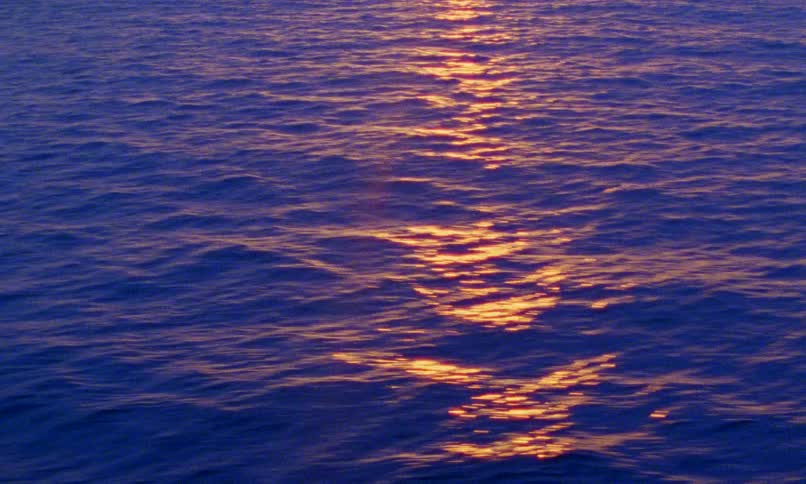 The golden light of a setting sun reflects on the deep blue, rippling surface of the ocean
