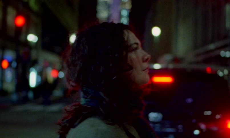 A woman with curly hair walks through a busy city at night as traffic and lights blur in the background