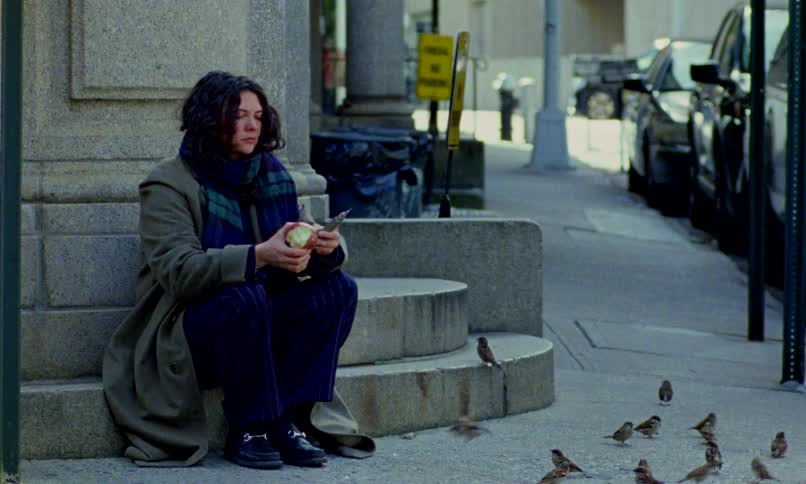 A pensive person with long hair peels an apple on a city sidewalk, surrounded by sparrows and pigeons