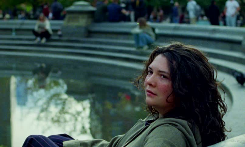 A troubled young woman with a bruised face sits by a fountain in a public park, looking sadly into the camera