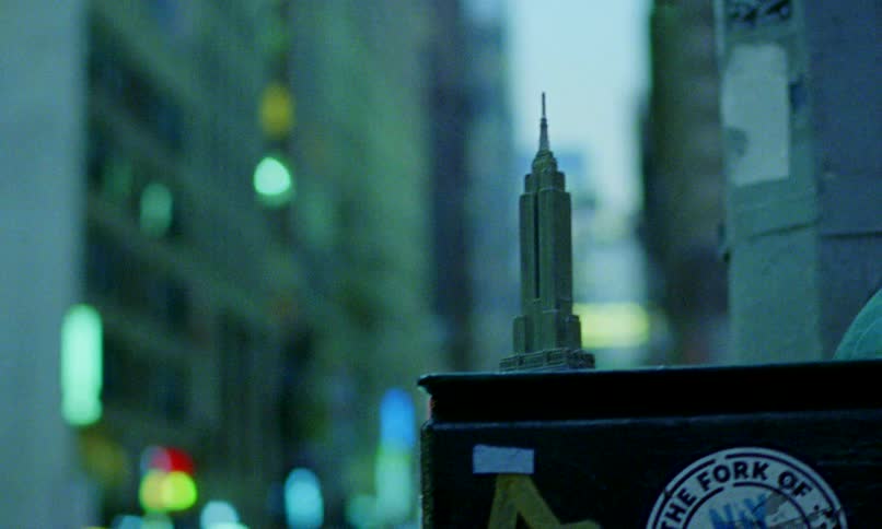 A woman places a souvenir Empire State Building on a street post before pensively watching the blurry city lights of New York City at night