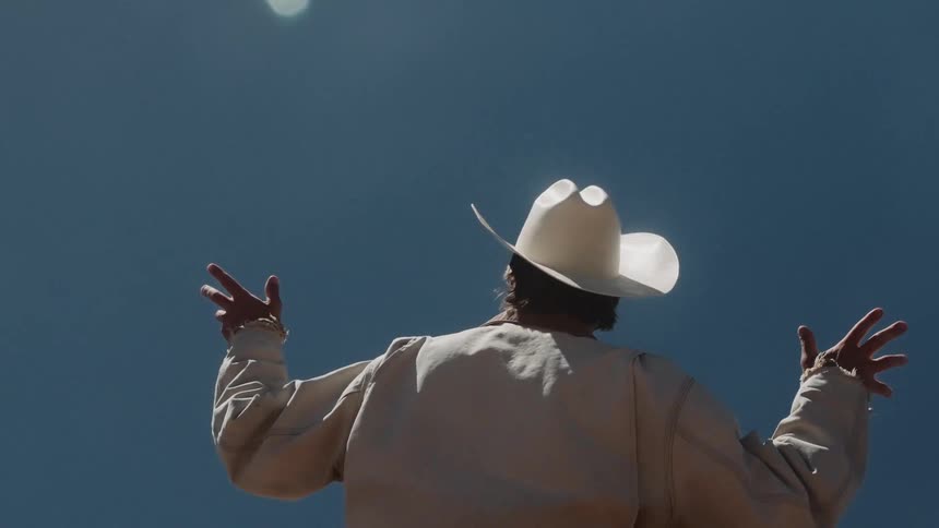 A figure in a white cowboy hat slowly raises their arms towards the sun against a clear blue sky in a spiritual gesture