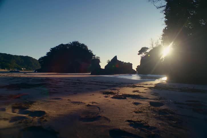 The sun shines brightly over a tranquil beach cove with silhouetted rock formations and footprints in the sand