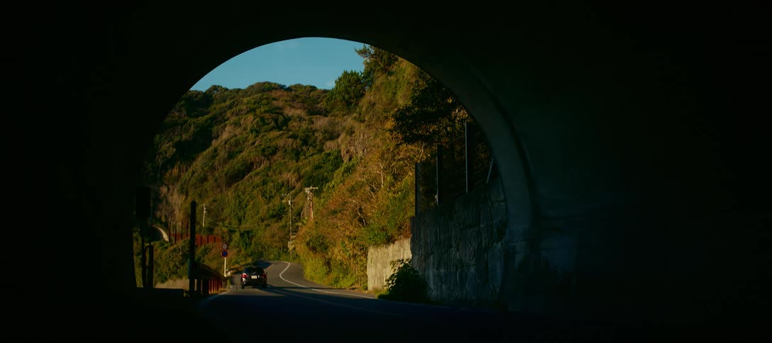 A silhouetted car exits a dark tunnel onto a sunny, winding road along a green hillside