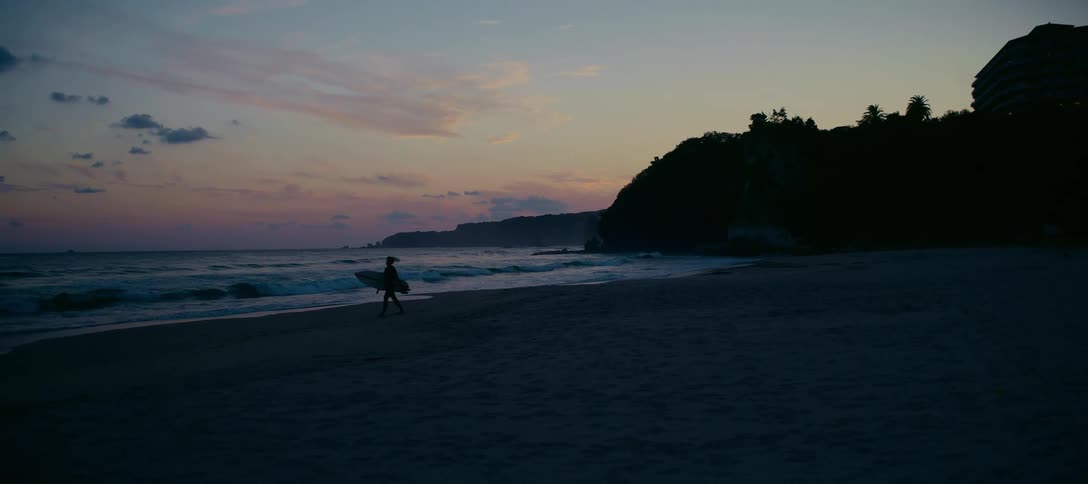 A silhouetted surfer with a surfboard walks along the beach next to the ocean at dusk