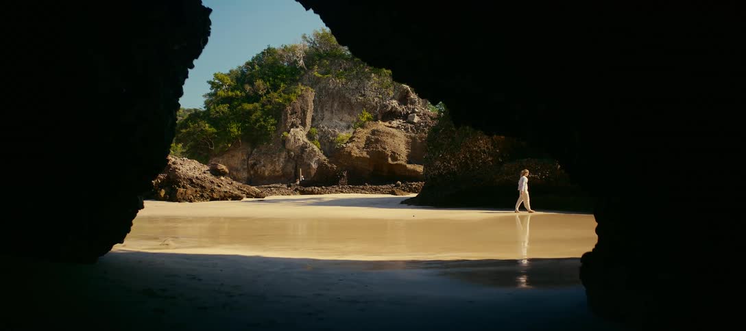 A lone woman walks along a secluded tropical beach, framed by the silhouette of a dark seaside cave
