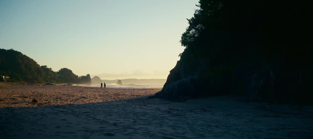 A cinematic wide shot shows the silhouette of two people walking along a serene beach during a golden sunset