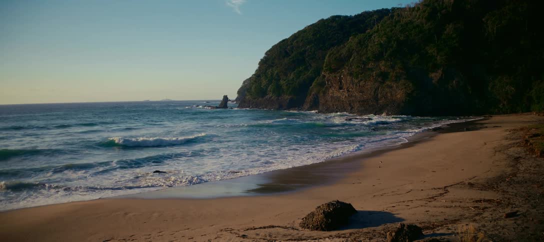 A serene, wide shot of a secluded sandy beach with gentle waves lapping the shore next to a lush, green cliffside during golden hour