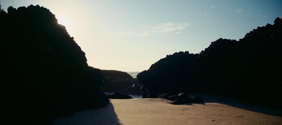 A powerful ocean wave crashes against silhouetted rocks in slow motion as seen from a sandy cove at sunrise