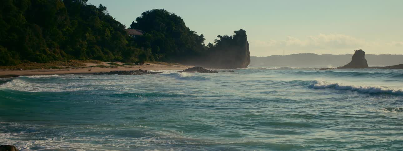 Slow motion shot of ocean waves rolling onto a misty shoreline with rocky cliffs in the warm morning light