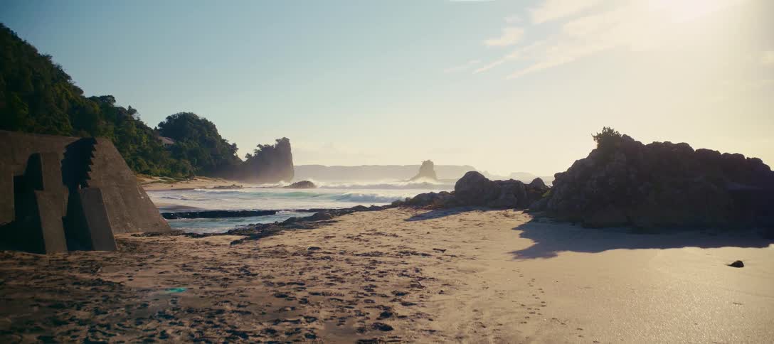 A serene coastal landscape features ocean waves gently breaking on a sandy beach surrounded by rocky cliffs on a bright, sunny day