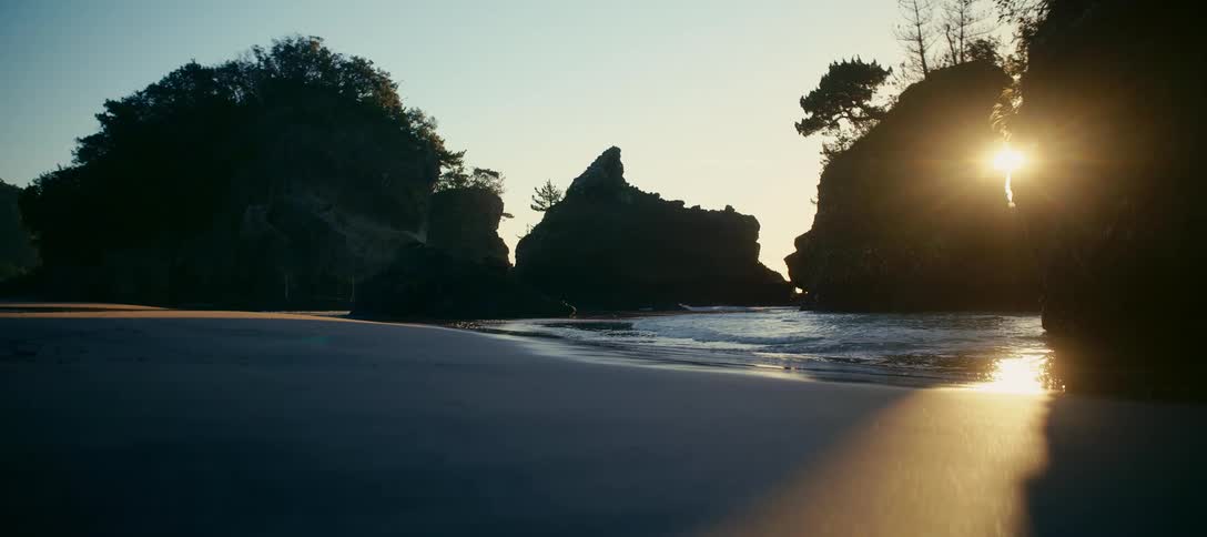 Golden sunset light reflects on the calm ocean waves lapping at a sandy beach surrounded by silhouetted rock formations