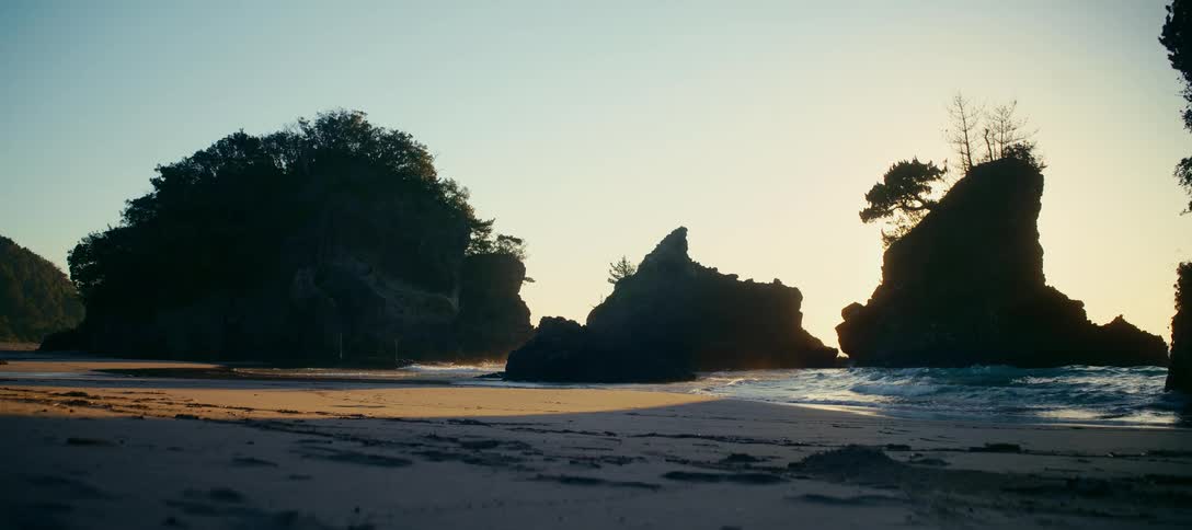 Silhouetted sea stacks stand against a golden sunrise as gentle waves wash onto the sandy shore