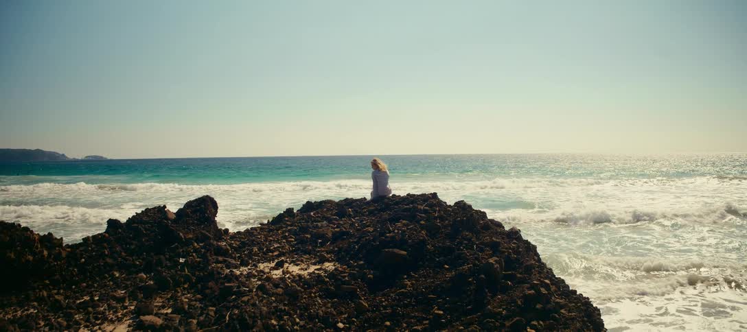 A solitary woman sits on a rugged rock formation, watching the turquoise ocean waves crash against the shore