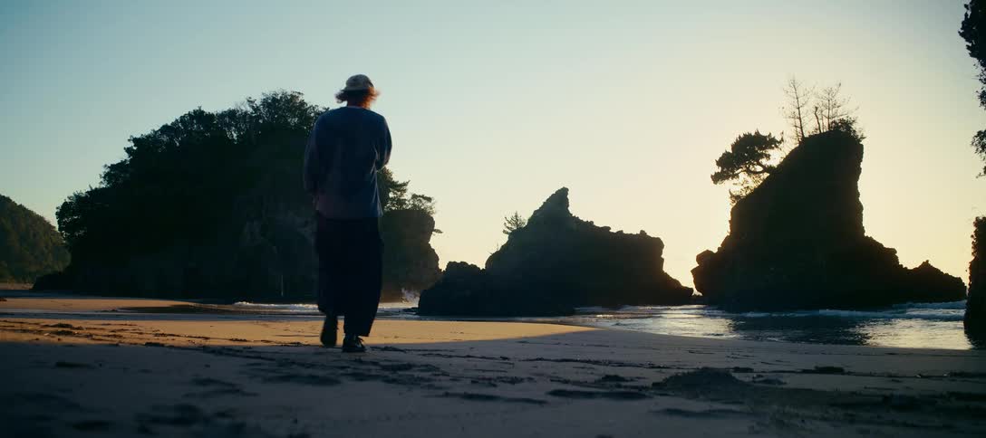 A lone figure walks along a sandy beach toward silhouetted sea stacks during a peaceful golden hour sunset