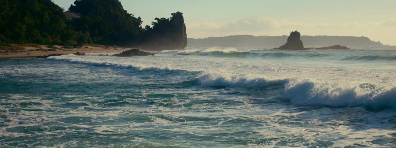 Slow motion capture of ocean waves crashing onto a tranquil, misty coastline with lush, tree-covered rock formations