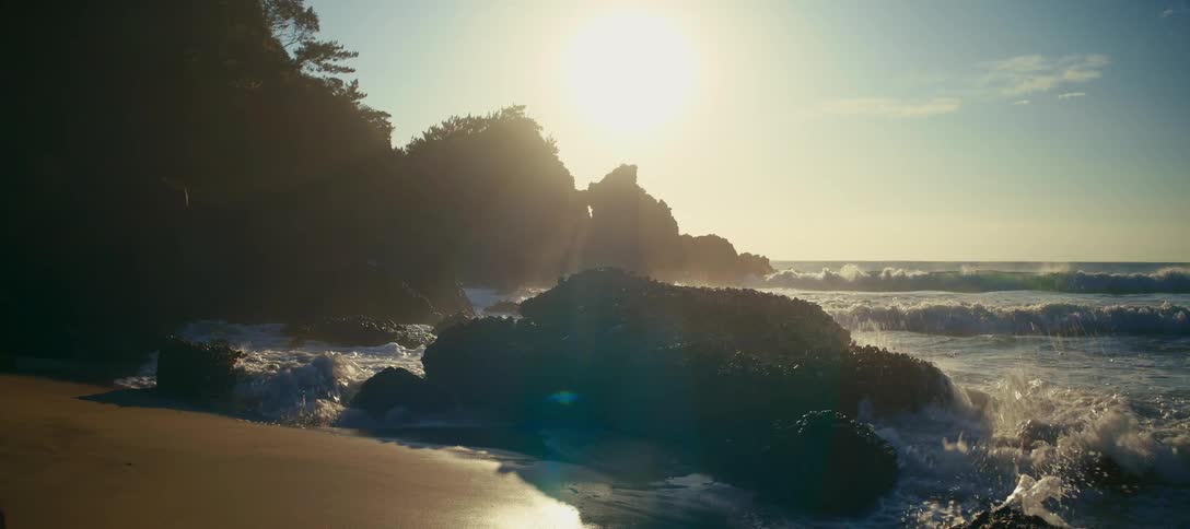 The bright sun shines over a rocky coastline as ocean waves crash onto the shore in beautiful slow motion