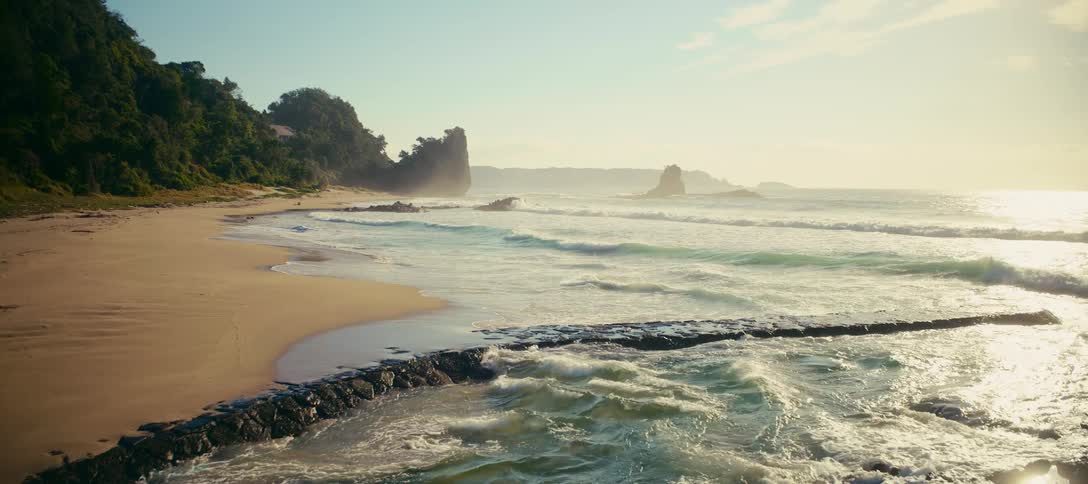 Gentle ocean waves wash over a rocky breakwater along a secluded sandy beach during a beautiful golden sunrise