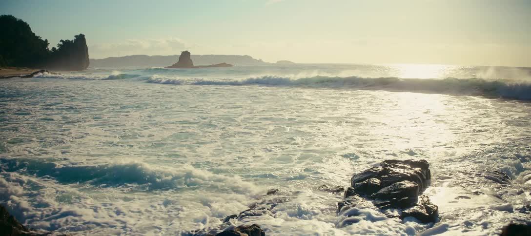 Sun-drenched ocean waves roll and crash onto a rocky coastline in dramatic slow motion