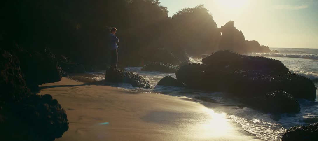 A lone woman stands on a rock, taking in the serene view as ocean waves crash on the sandy shore during a beautiful golden sunrise