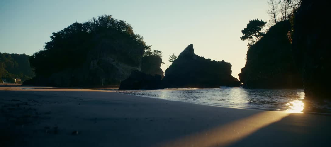 A serene view of a sandy beach cove with silhouetted rock formations as gentle waves wash ashore during a golden sunset