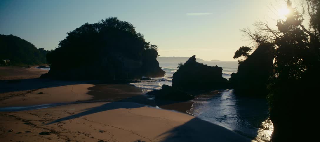The bright sun rises over a serene sandy beach cove with large silhouetted rock formations