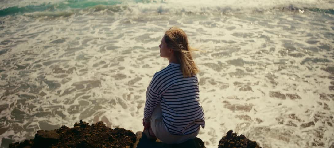 A contemplative woman with windblown hair sits on a rocky shoreline, peacefully watching the sunlit ocean waves