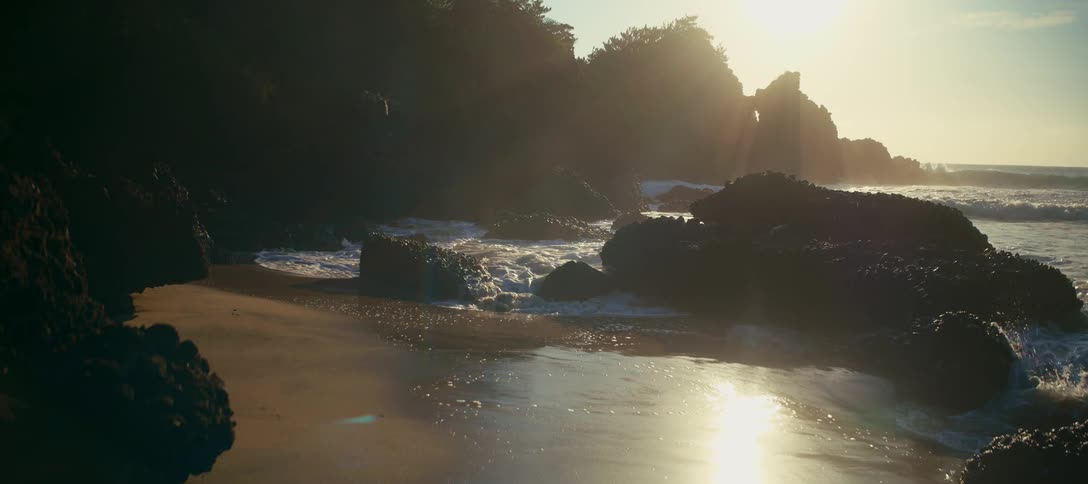 Cinematic slow motion of gentle ocean waves washing onto a rocky sand beach backlit by the rising sun