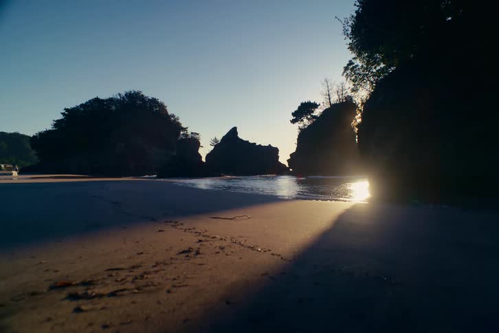 The morning sun shines between silhouetted rocks on a calm and sandy beach as waves gently lap the shore