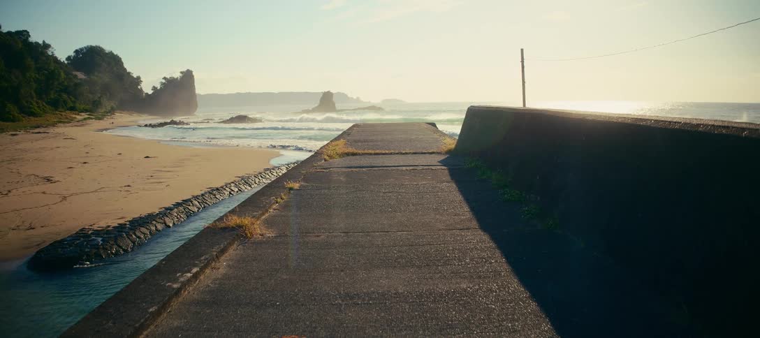A scenic view from a concrete pier shows the gentle waves of the ocean rolling onto a sandy beach at sunrise