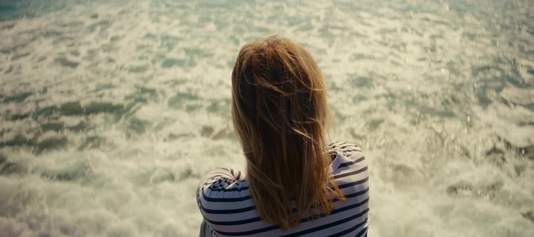 Slow motion shot of a woman with her back to the camera, gazing at the sparkling ocean and shielding her eyes from the sun